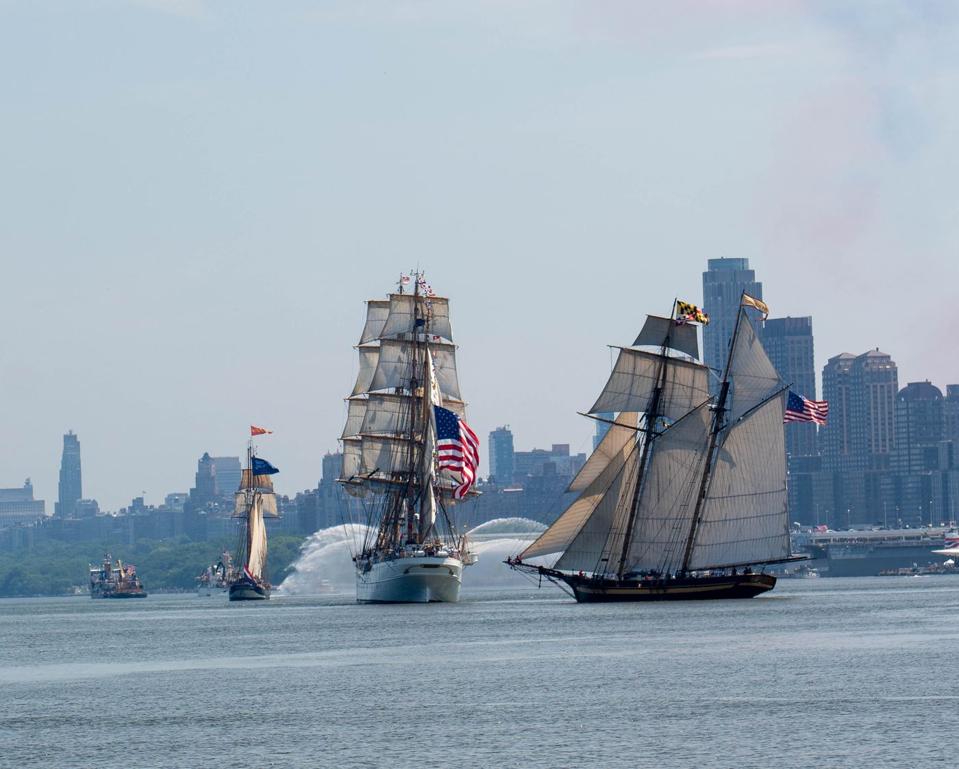 Three tall ships and a fireboat on the Hudson River with the Manhattan skyline behind them